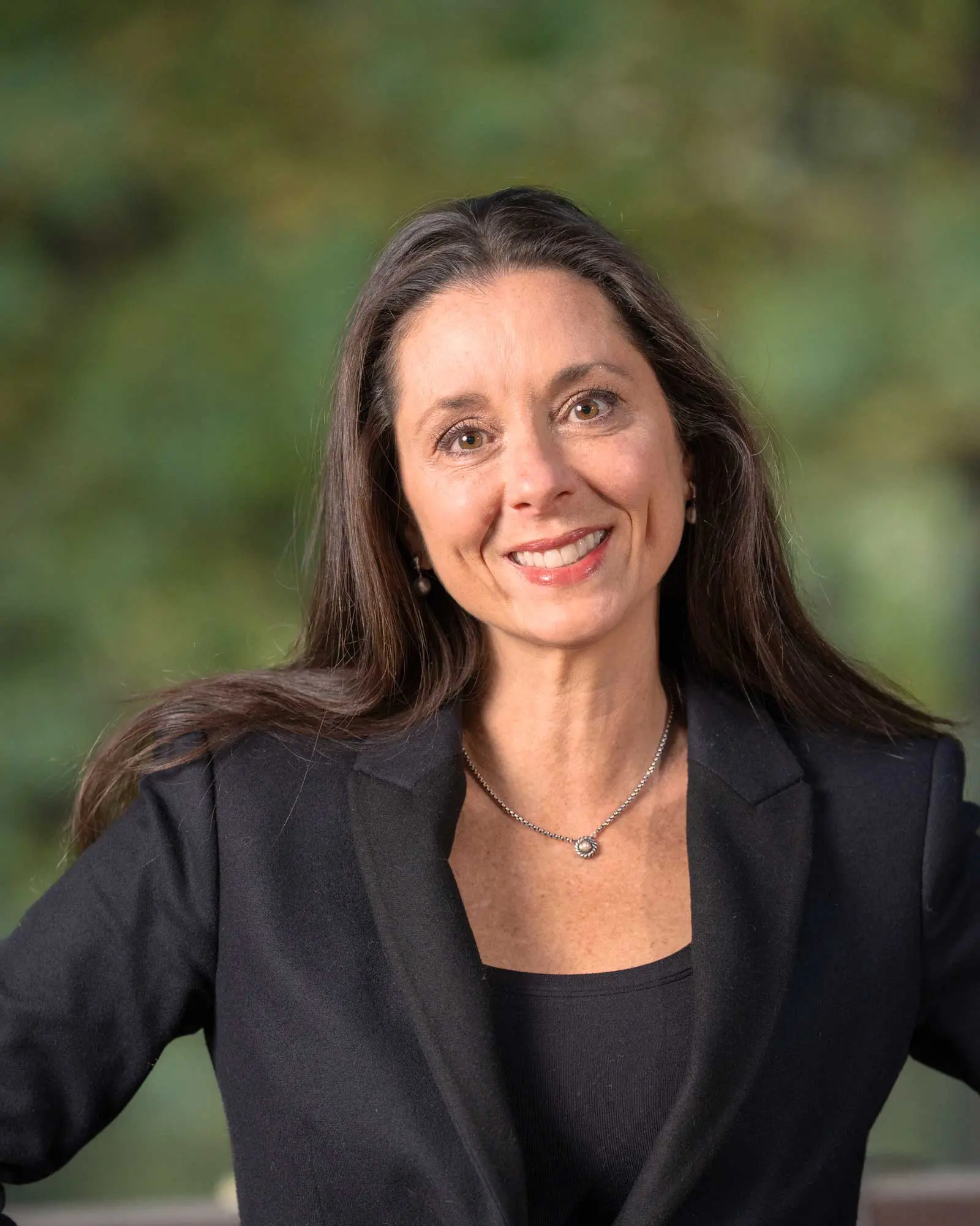 Woman with long brown hair wearing a black blazer and necklace, smiling at the camera with a blurred green background.