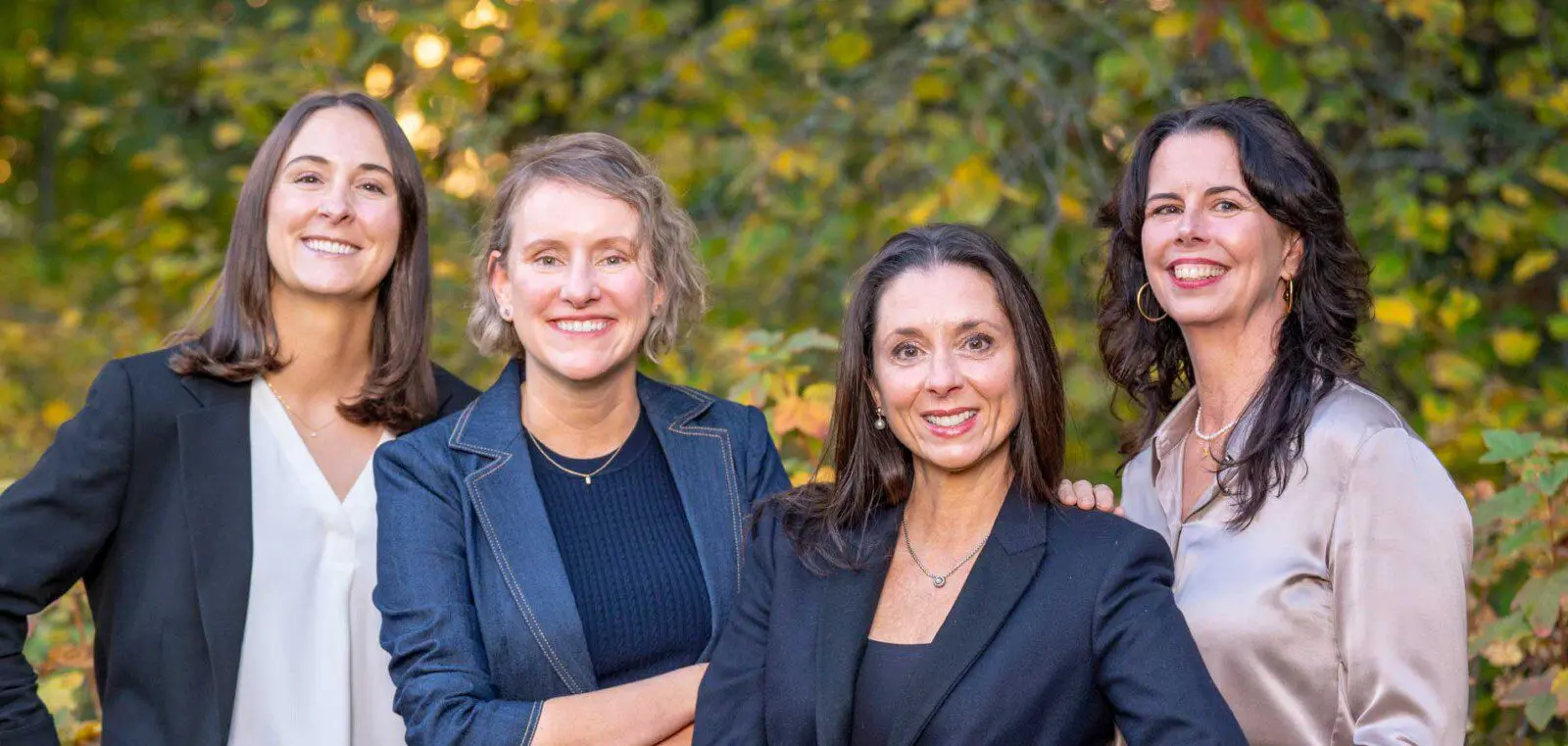 Four women standing outdoors in front of greenery, smiling and dressed in business casual attire.