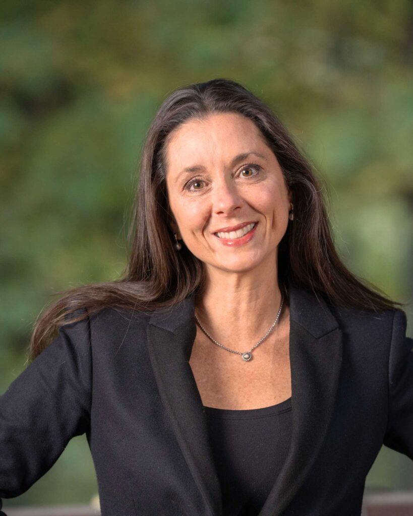 Woman with long brown hair wearing a black blazer and necklace, smiling at the camera with a blurred green background.