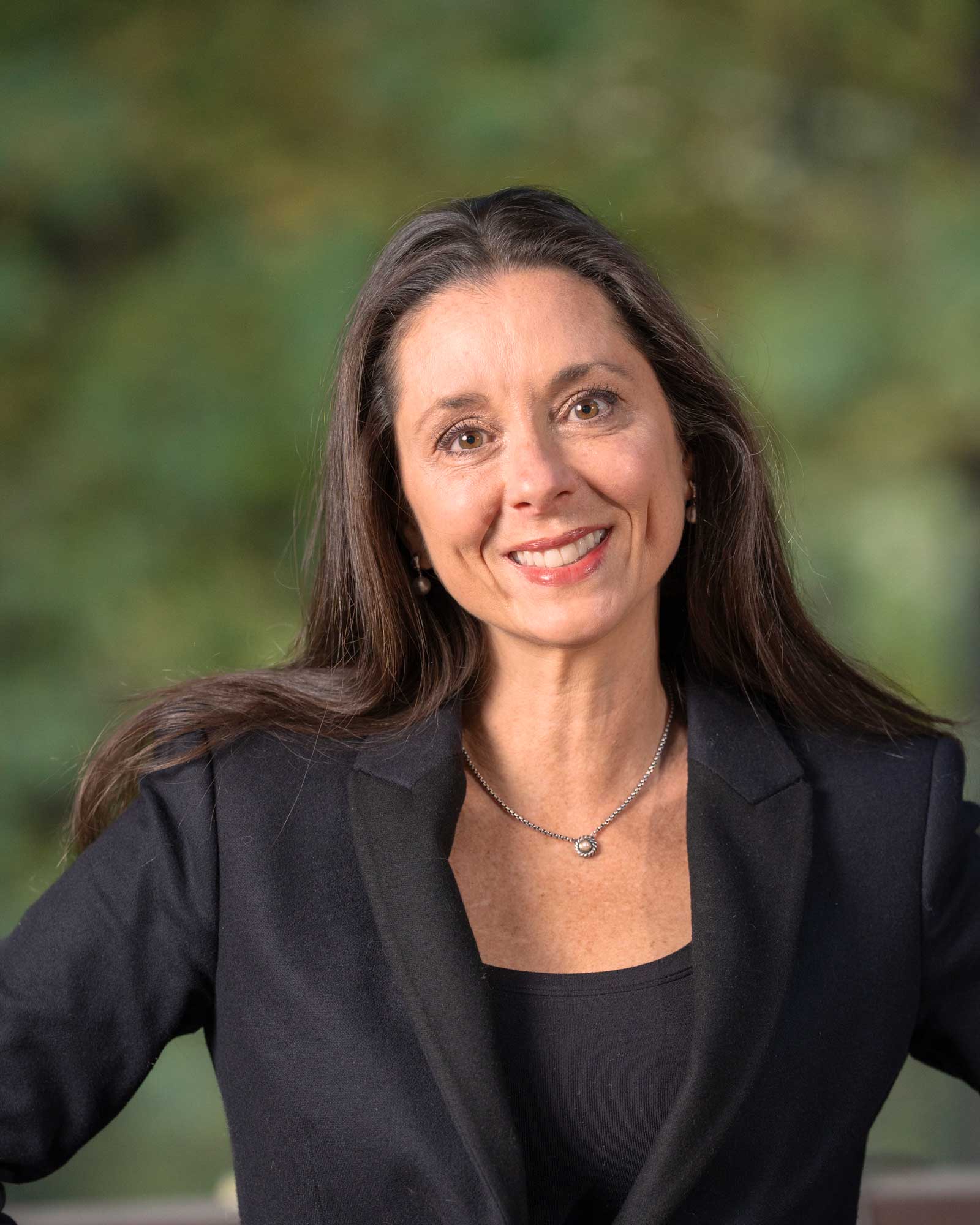 Woman with long brown hair wearing a black blazer and necklace, smiling at the camera with a blurred green background.