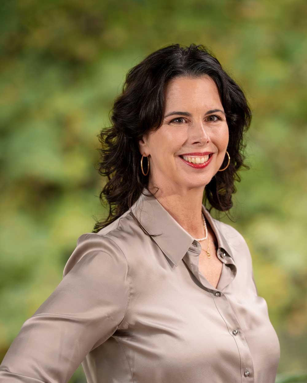 Woman with dark wavy hair wearing a beige satin blouse and gold hoop earrings, smiling and standing in front of a blurred green background.